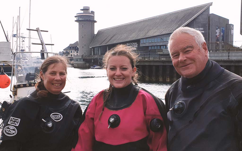 three divers in dry suits smiling at a pier during a dive trip
