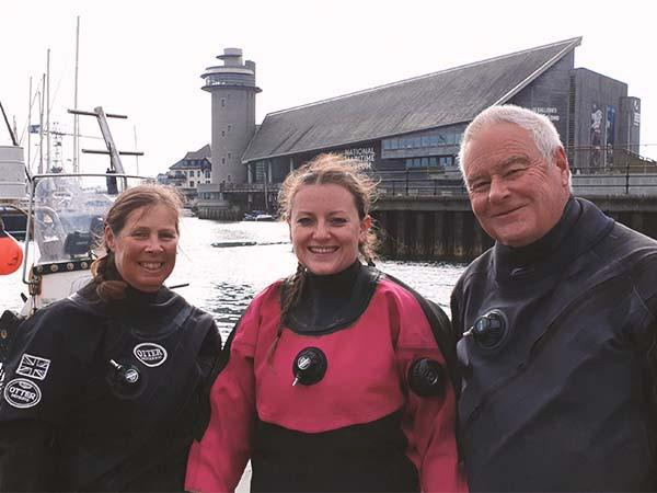 three divers in dry suits smiling at a pier during a dive trip
