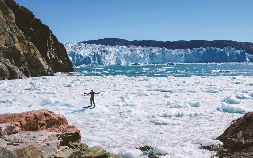 Andy Torbet in a drysuit standing with arms out on ice near a glacier