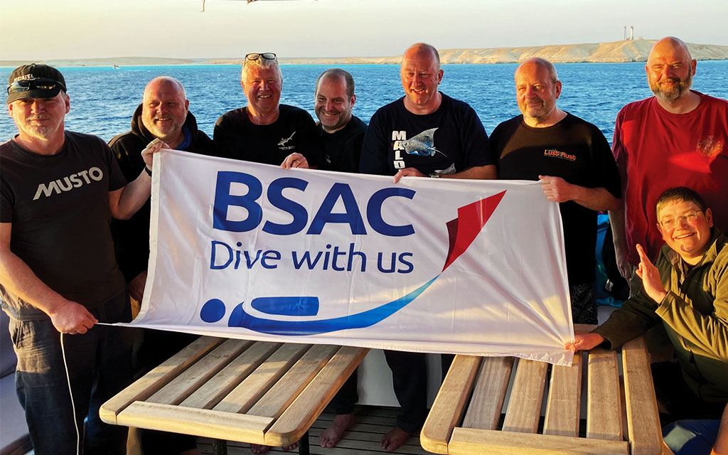 First Class Diver candidates holding the BSAC banner, blue ocean in the background with a landmass in the distance with a lighthouse