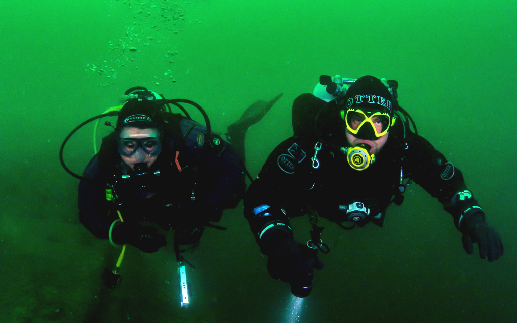 Two scuba divers, mid-dive, illuminated by the flash from the camera. Green, murky waters in the background.