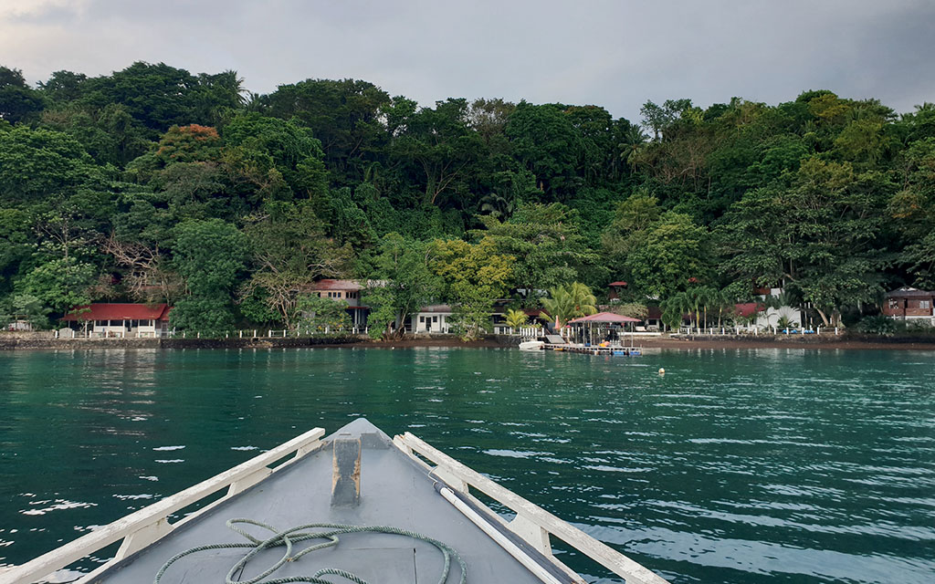 A boat approaches a resort on a densely forested Indonesian coast over emerald green waters 