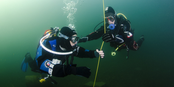 Two scuba divers in murky blue waters ascend on a yellow line