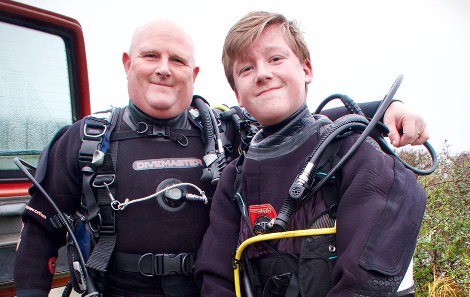 A man and a teenage boy stand side by side in scuba kit, smiling at the camera